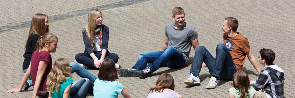 Eine weibliche Erwachsene spricht mit Schülerinnen und Schülern im Pausenhof im Sitzkreis