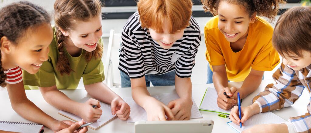 Junge Schülerinnen und Schüler lösen gemeinsam an einem Tisch unter Zuhilfenahme eines Tablets Aufgaben.